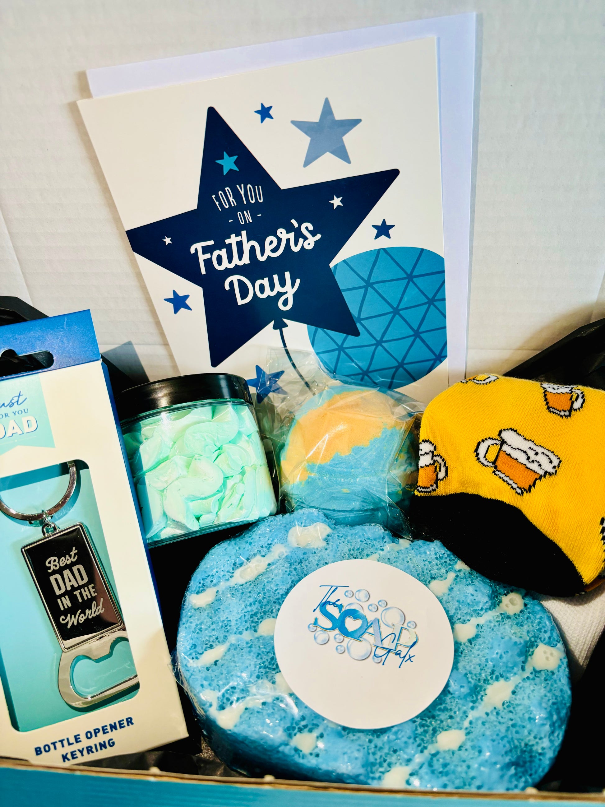 A Father's Day gift set featuring aftershave-inspired scents, a card, bottle opener keyring, bath bombs, socks with beer mug design, and mint candies in a tin is available as the **Men’s Father's Day Skincare Gift Set Door To Door** by **The Soap Gal x**.
