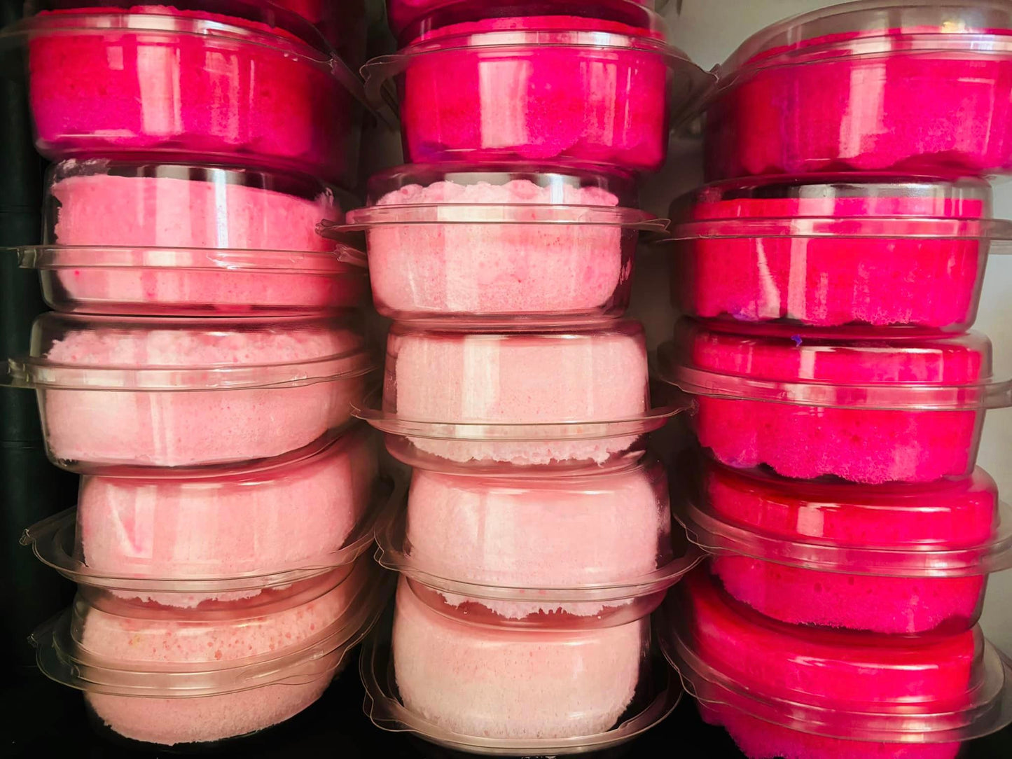 Stacks of clear plastic containers filled with The Soap Gal x's pink and magenta Random Scented Exfoliating Soap Sponges, available with a special BUY 2 GET 65% OFF THE 3RD SOAP SPONGE offer, are arranged in three columns.
