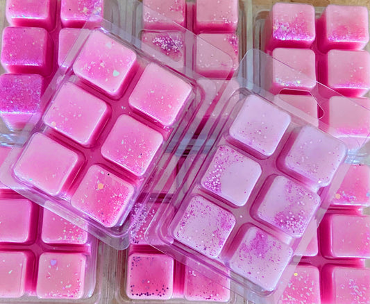 Several packages of pink wax melts with glitter are stacked and arranged in a pattern. Each Wax Melt Gift Set 4 or 6 from The Soap Gal x contains six individual melts housed in PET recycled plastic clamshells, offering an eco-friendly touch at a discounted price.