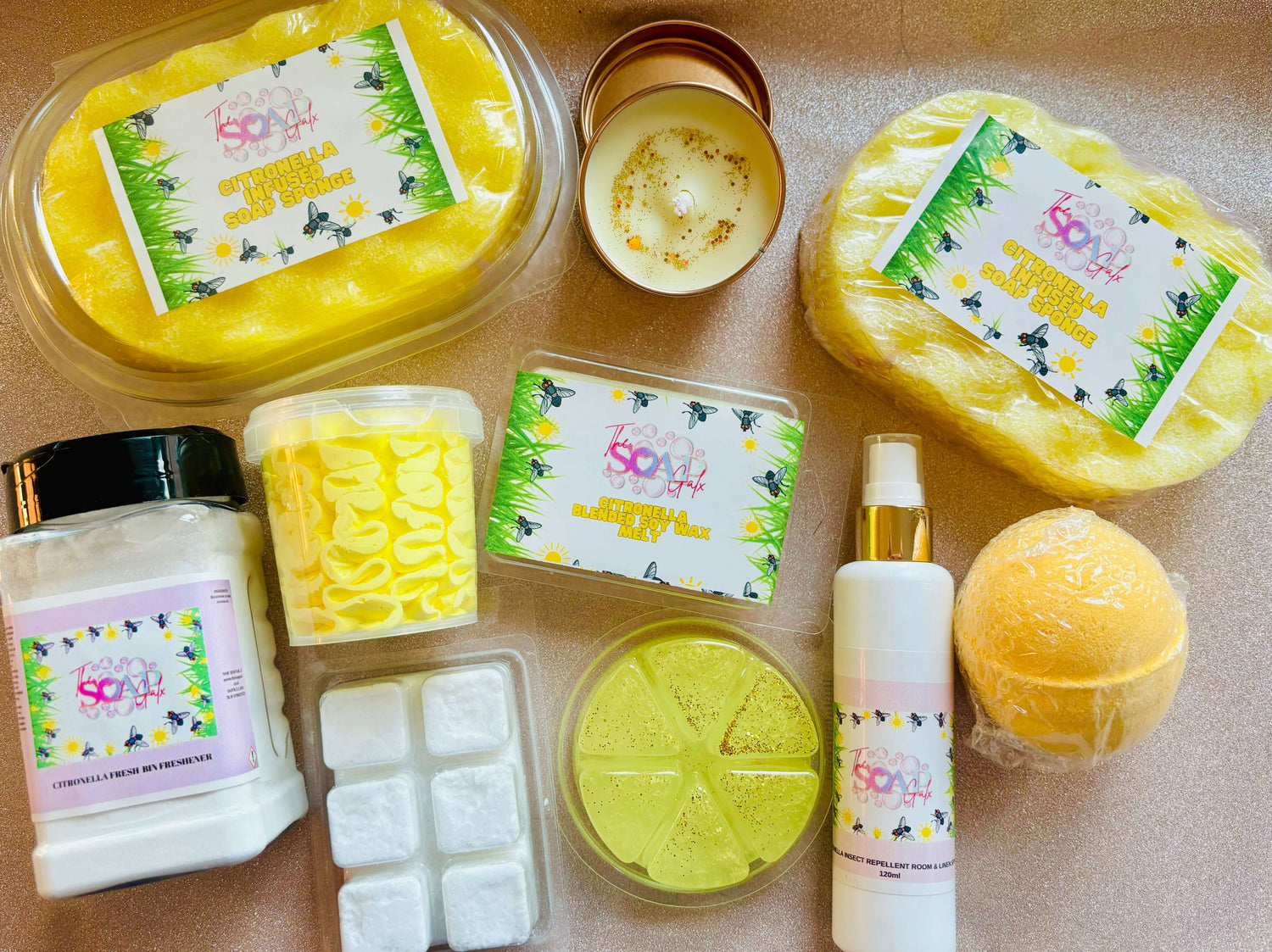 A flat lay of various bath products, including soap bars, a candle featuring The Soap Gal x Citronella Insect Repellent Skincare & Home Fragrance Bundle, lotion, salt, bath fizzies, and a bath bomb, all with colorful packaging.